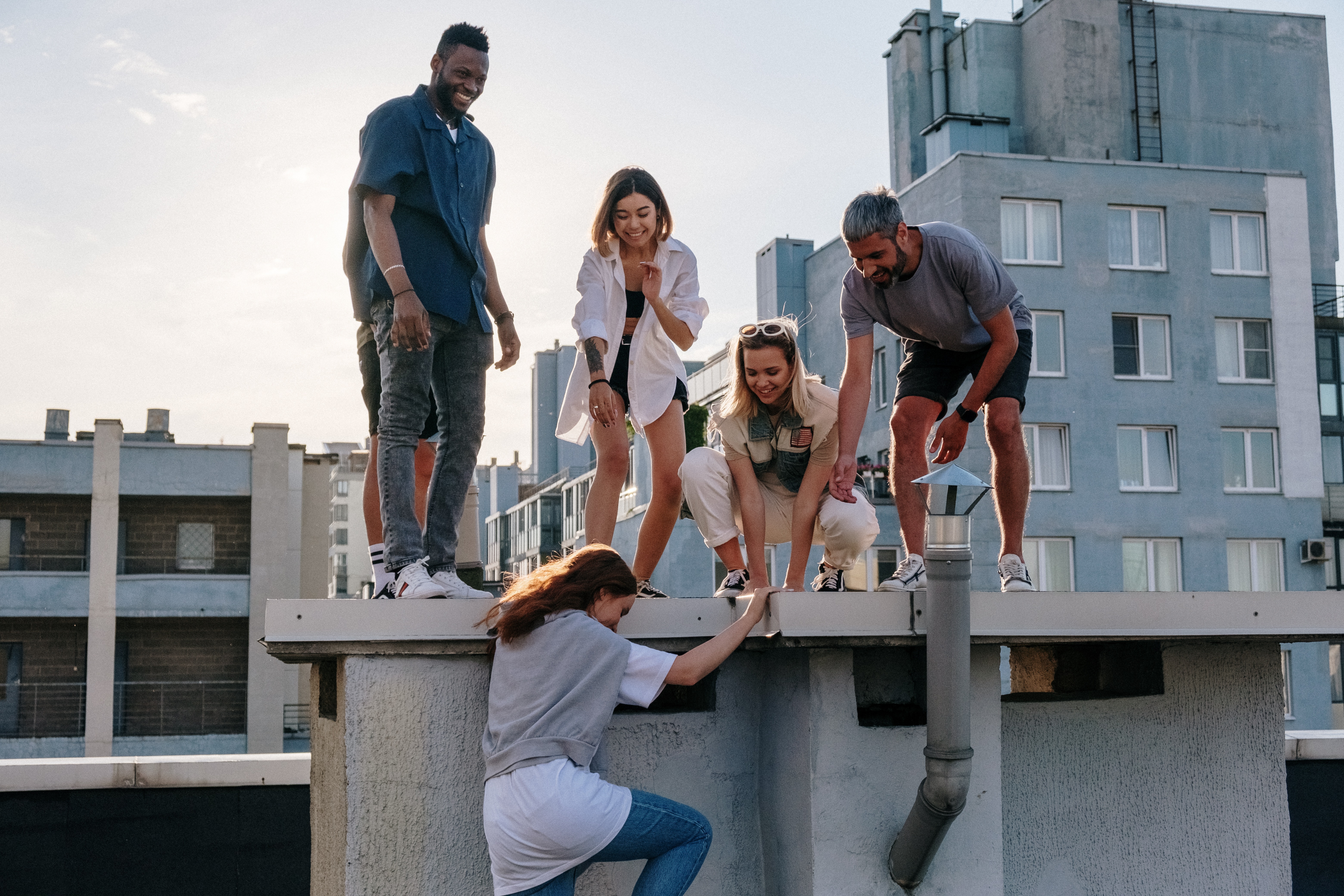 A group of 5 multi ethnic and attractive young men and women. They are all helping one climb to where they are on the roof of a building.