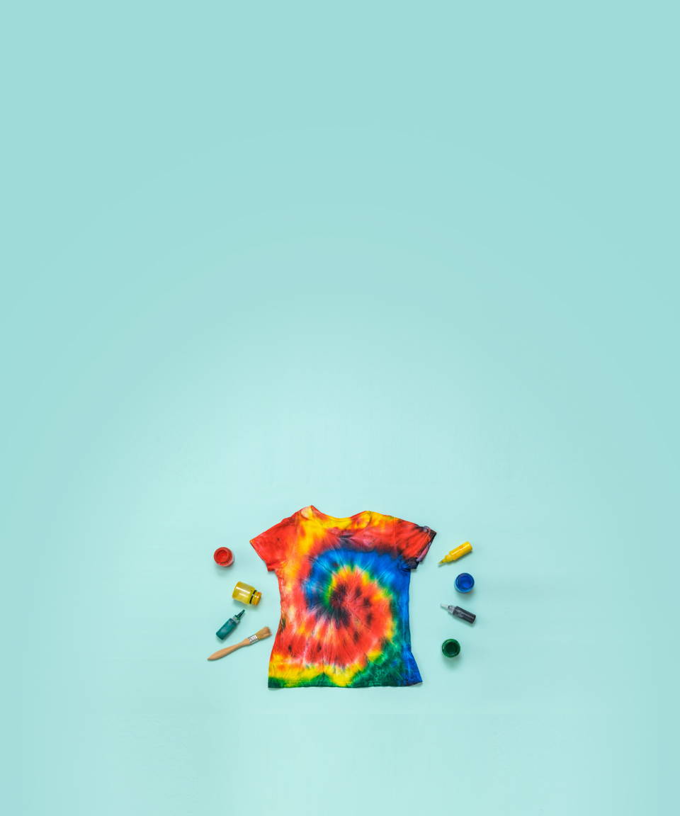A tie dyed t-shirt surrounded by colorful ink dropper bottles and a paint brush for Confetti's Virtual Tie Dye Workshop