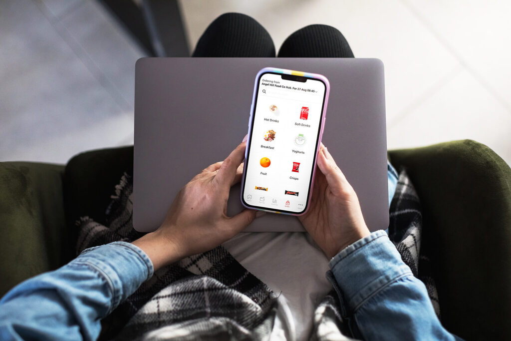 A person sits with a laptop on their lap, holding a smartphone displaying a grocery shopping app with categories like breakfast, fruits, yoghurts, and chips. The person is wearing a plaid blanket and denim shirt.