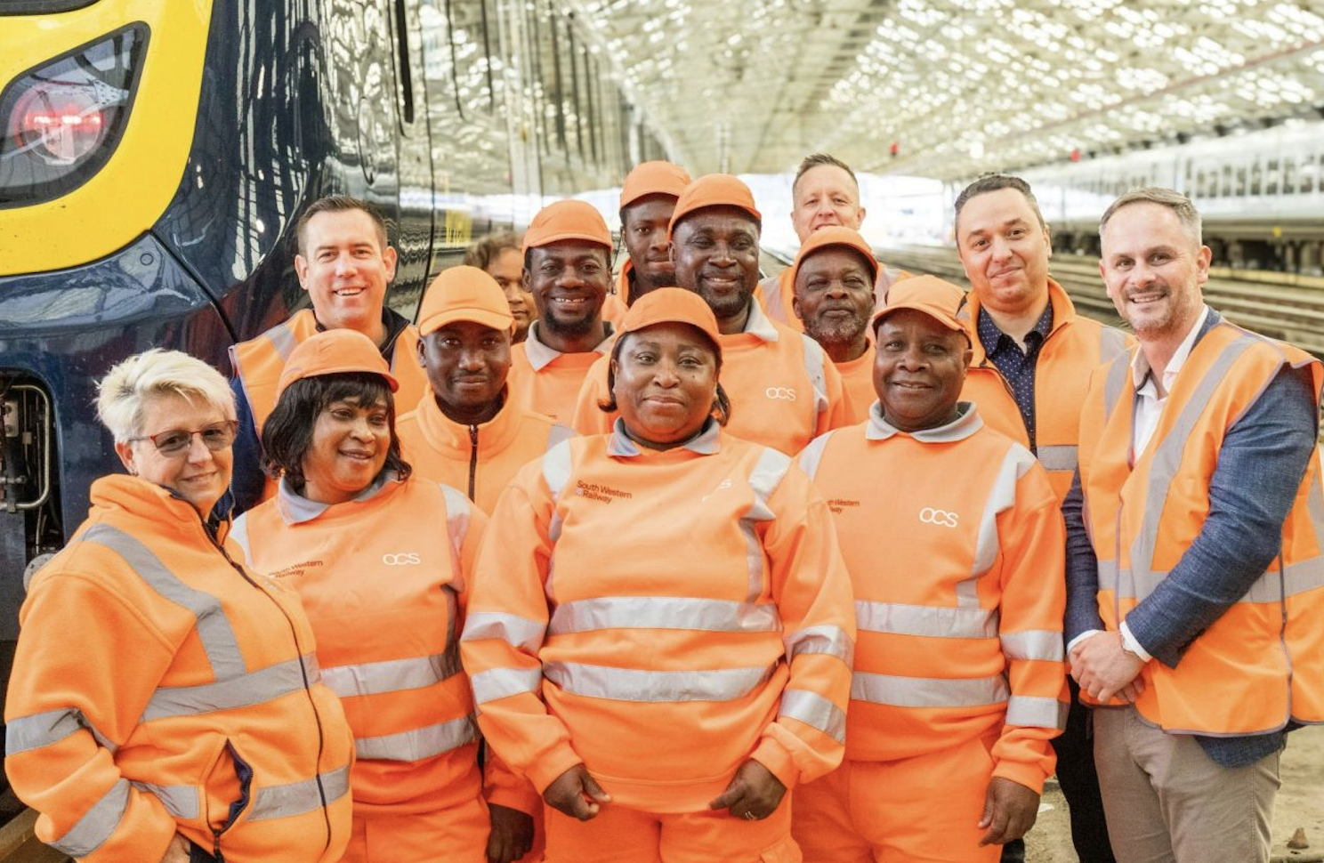 A group of rail cleaning workers in orange high-visibility uniforms and hats pose together on a train platform, standing beside a train inside a well-lit station. Some people wear safety vests and smile at the camera.