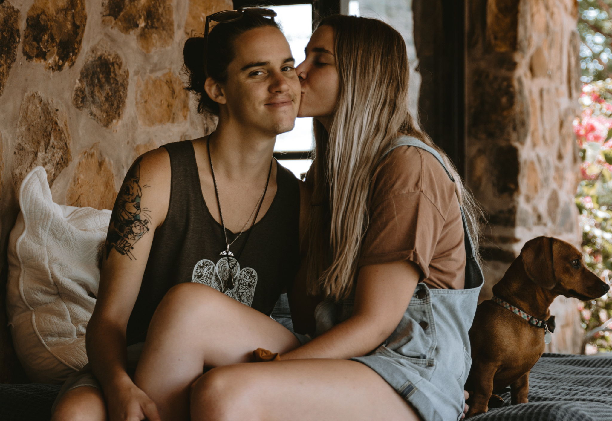 A woman sits on her side and on top of her partner kissing his cheek while he smiles and looks at the camera.
