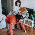 A man and woman work together to tidy up their living room. The man is shown lifting up a table, while the woman follows closely behind him, carrying a basket filled with pillows. The scene takes place in a cozy, well-lit flat, with an open doorway in the background revealing a bicycle parked in the hallway. Both individuals are dressed casually as they go about decluttering their living space.