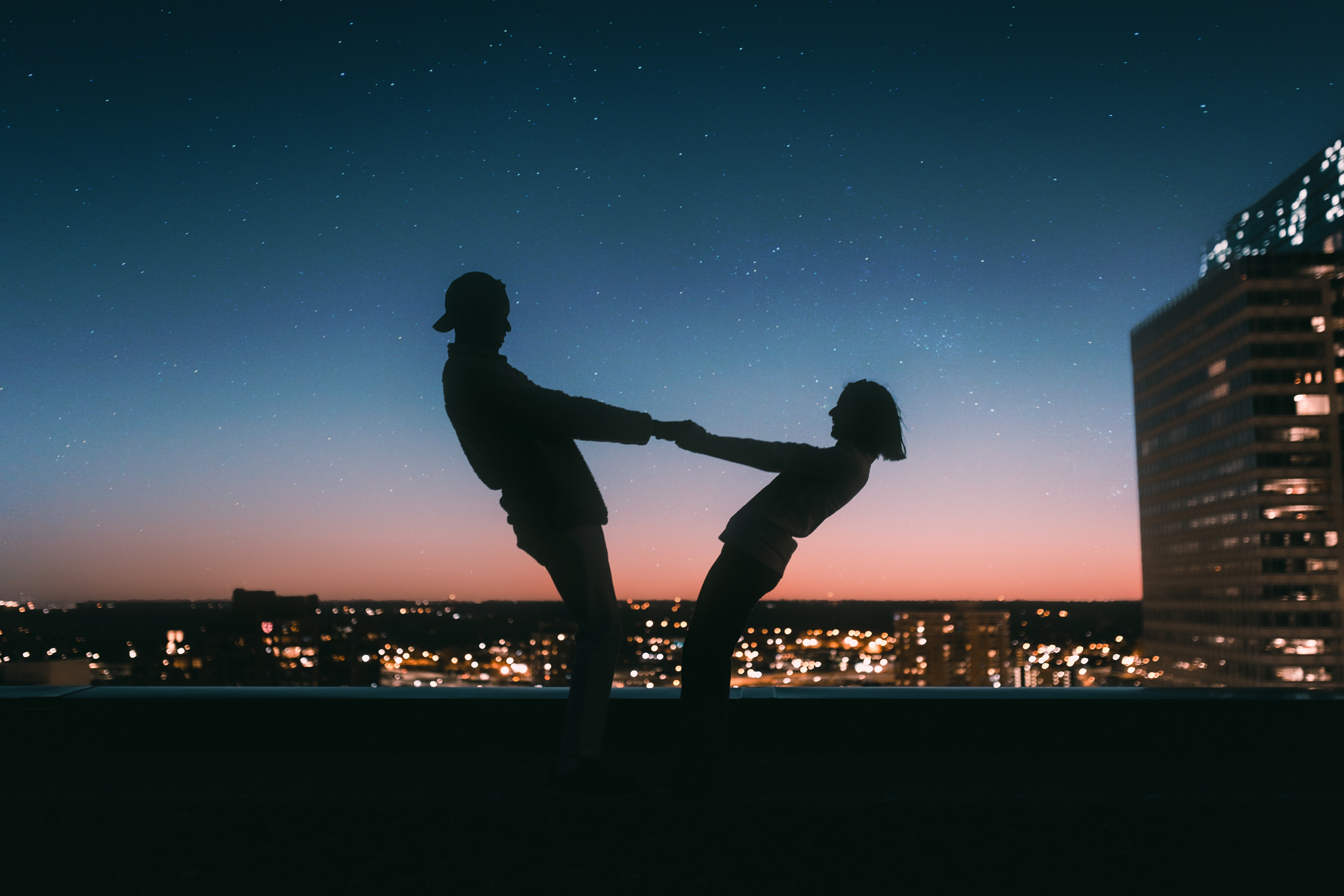 Image of a the silouhette of a man and a woman holding both hands together and leaning back, during dusk.