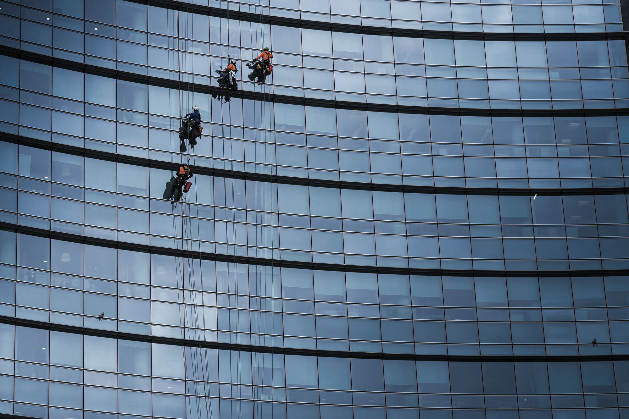 Three workers wearing safety gear clean windows while suspended on ropes outside a tall modern glass building, reflecting the city and sky.