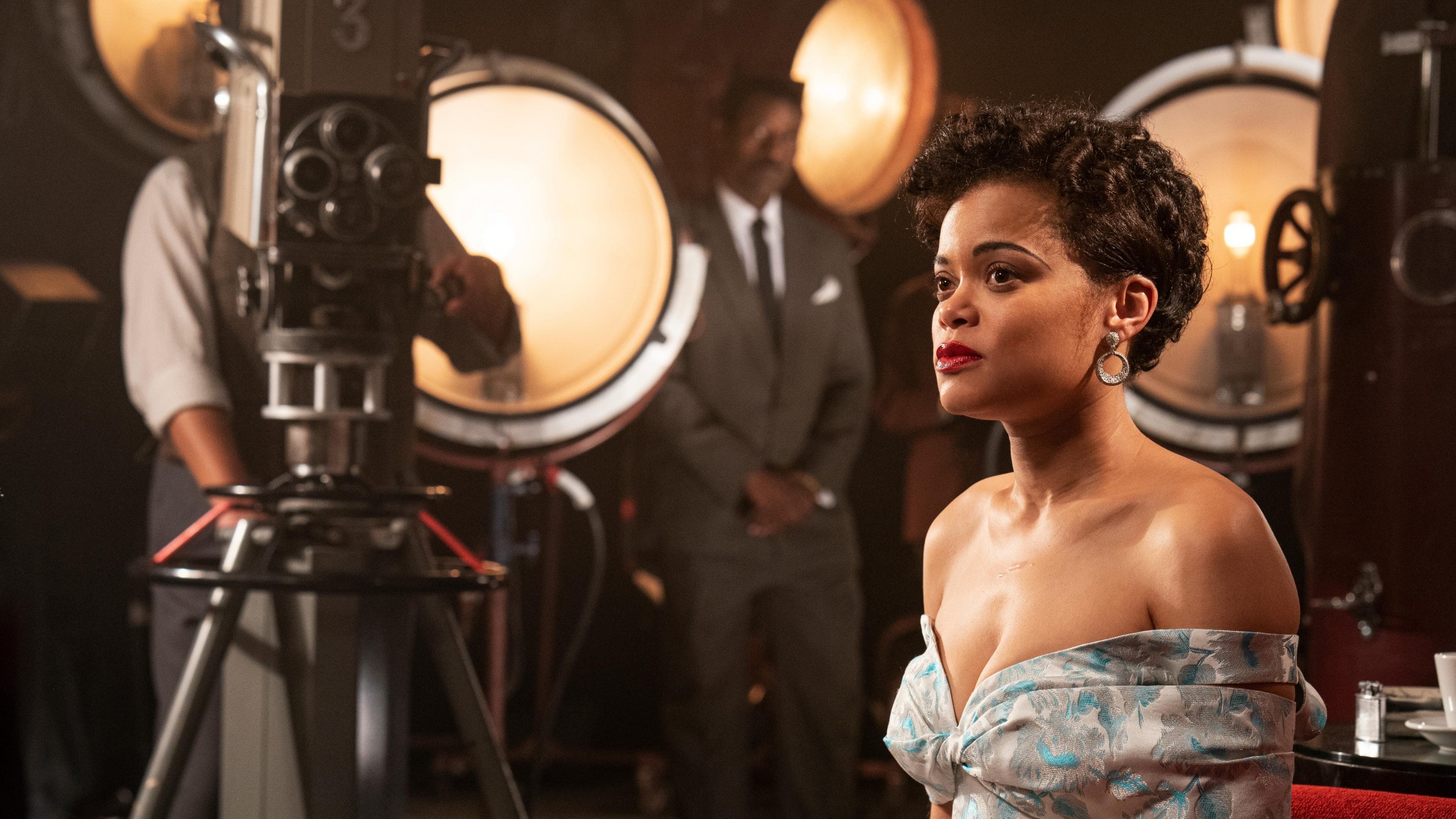 Image of Lady Day, sitting with cameras and lights all around her, she is looking at a distance and has a low cut dress.