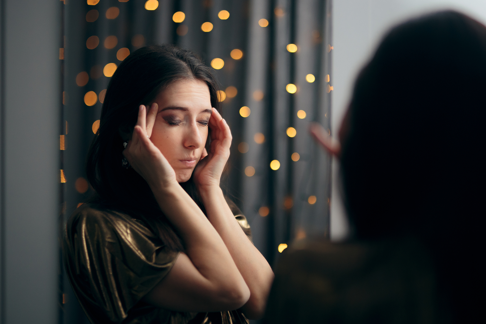 A young woman holds her temples and closes her eyes in the bathroom while at a holiday gathering.