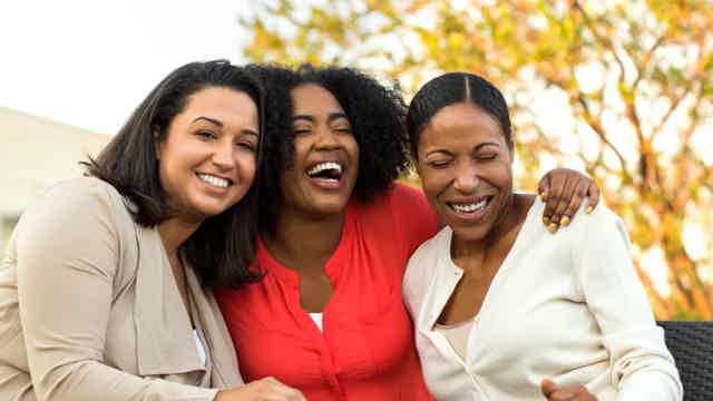Three women sit close together, smiling and laughing outdoors, arms around each other.