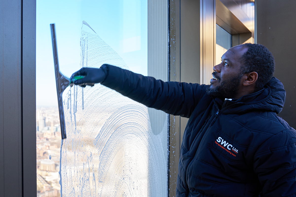 A person in a black SWC Ltd jacket uses a squeegee to clean the exterior window of a tall building, with a cityscape visible in the background.