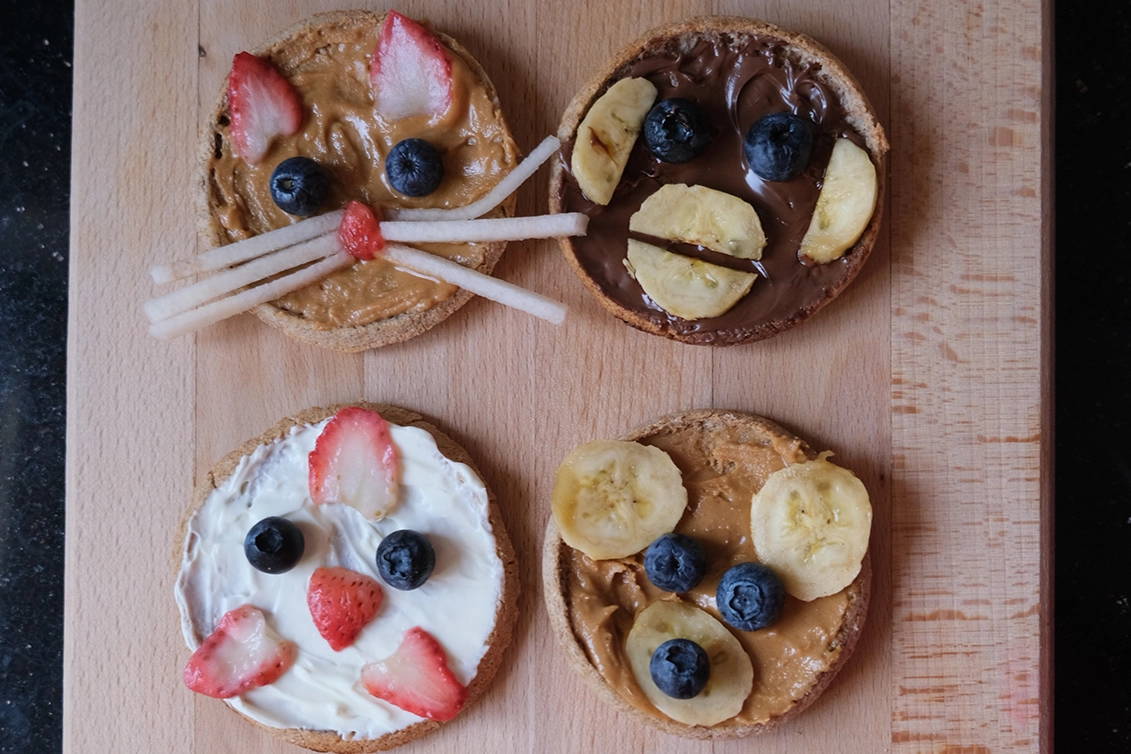 An overhead view of four Pacha English muffins decorated as animal faces using nut spreads, cream cheese, sliced strawberries, blueberries, and bananas, arranged on a wooden cutting board.
