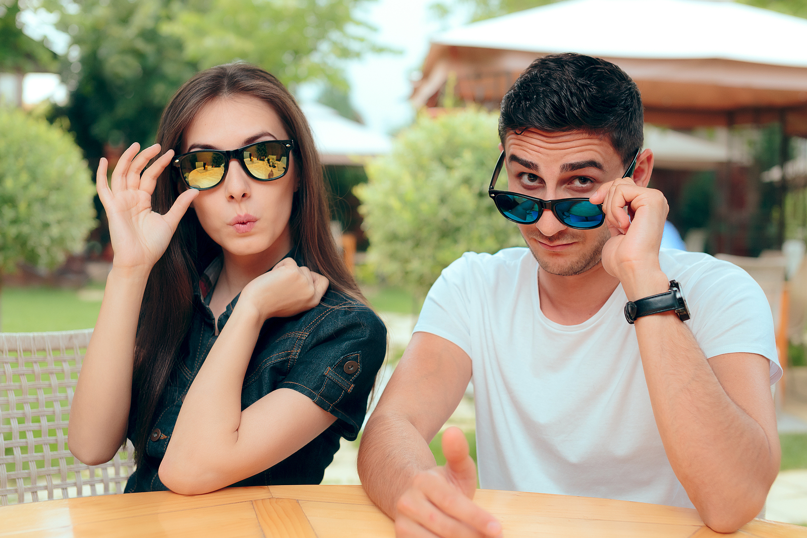 An attractive man and woman sitting together with sunglasses and a judgy expression on their faces looking at the camera.