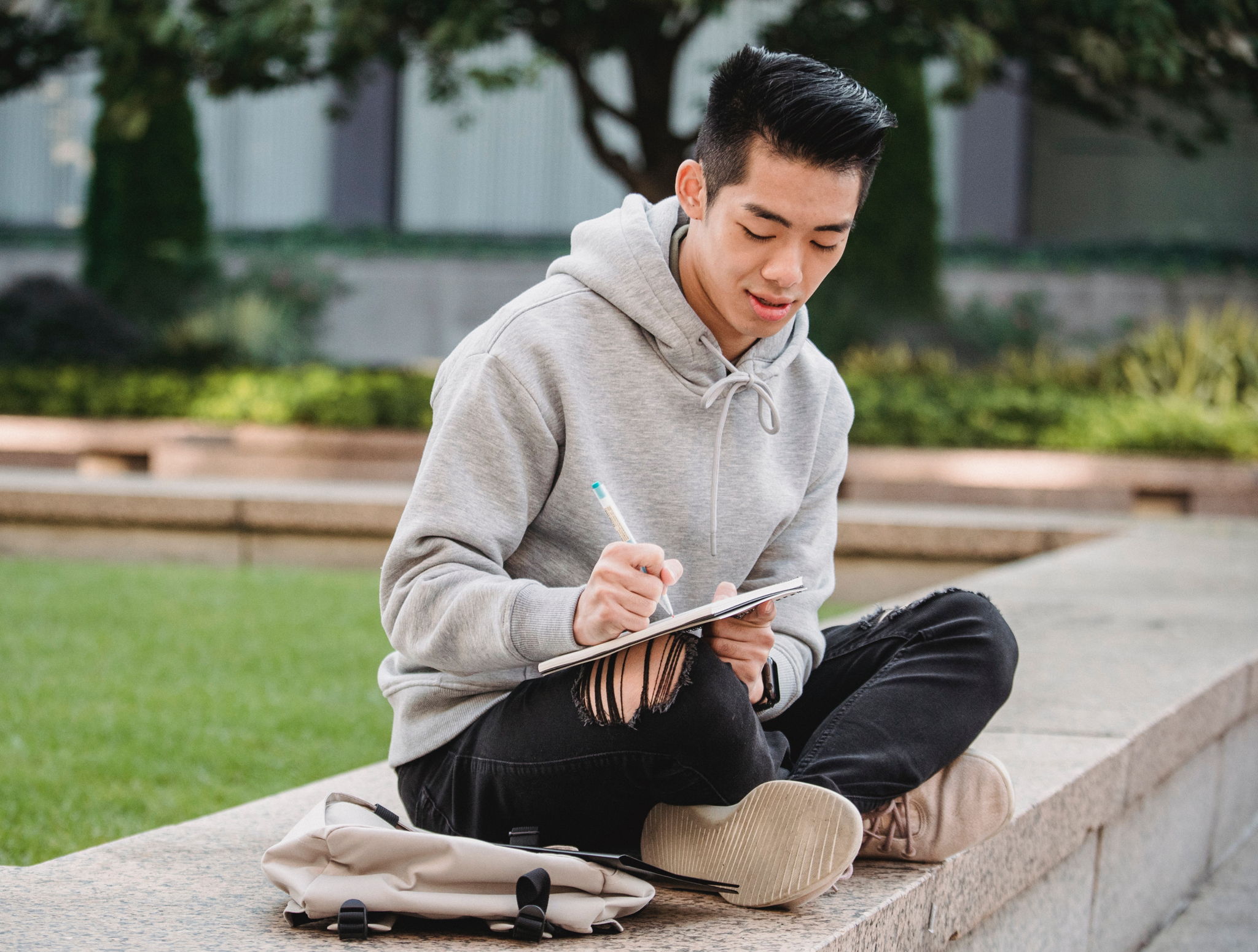 An asian american man sits cross legged drawing on a notepad outdoors.