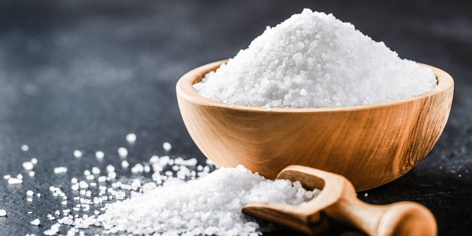 A small wooden bowl of coarse white salt, with matching wooden scoop, on a gray granite surface scattered with salt.