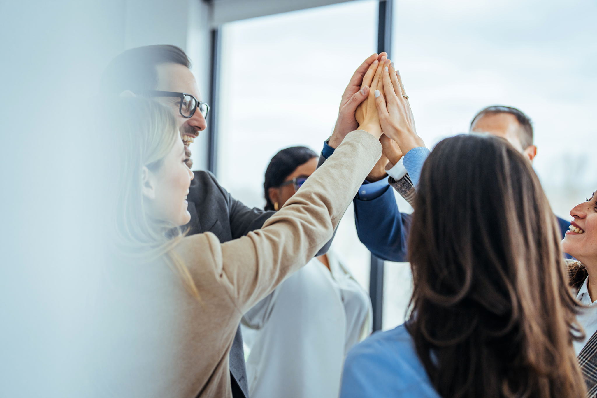 A group of professionals stand in a circle, smiling and giving a group high five, showing teamwork and celebration in a modern office setting with large windows.