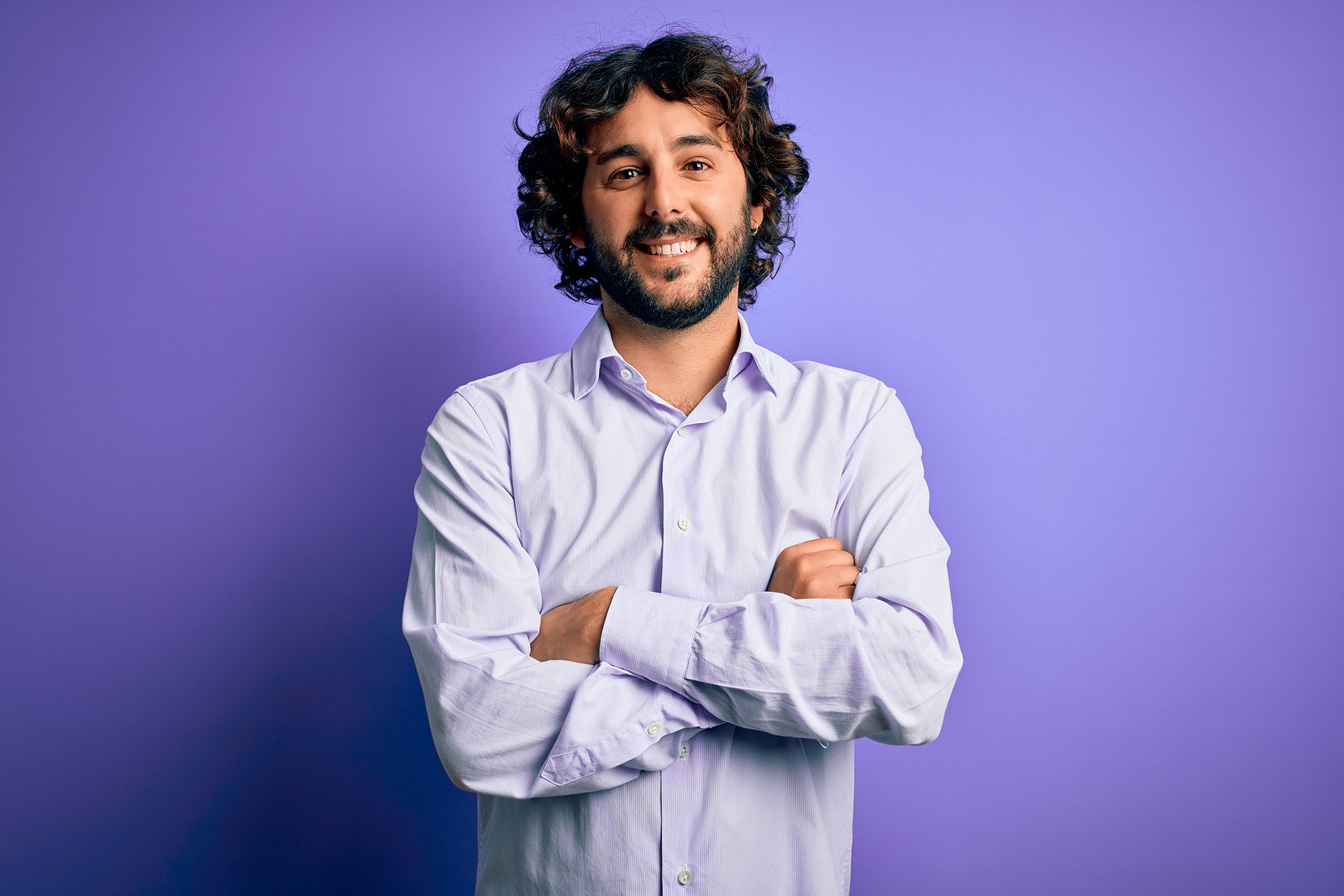 Young handsome business man with beard wearing shirt standing over purple background happy face smiling with crossed arms looking at the camera