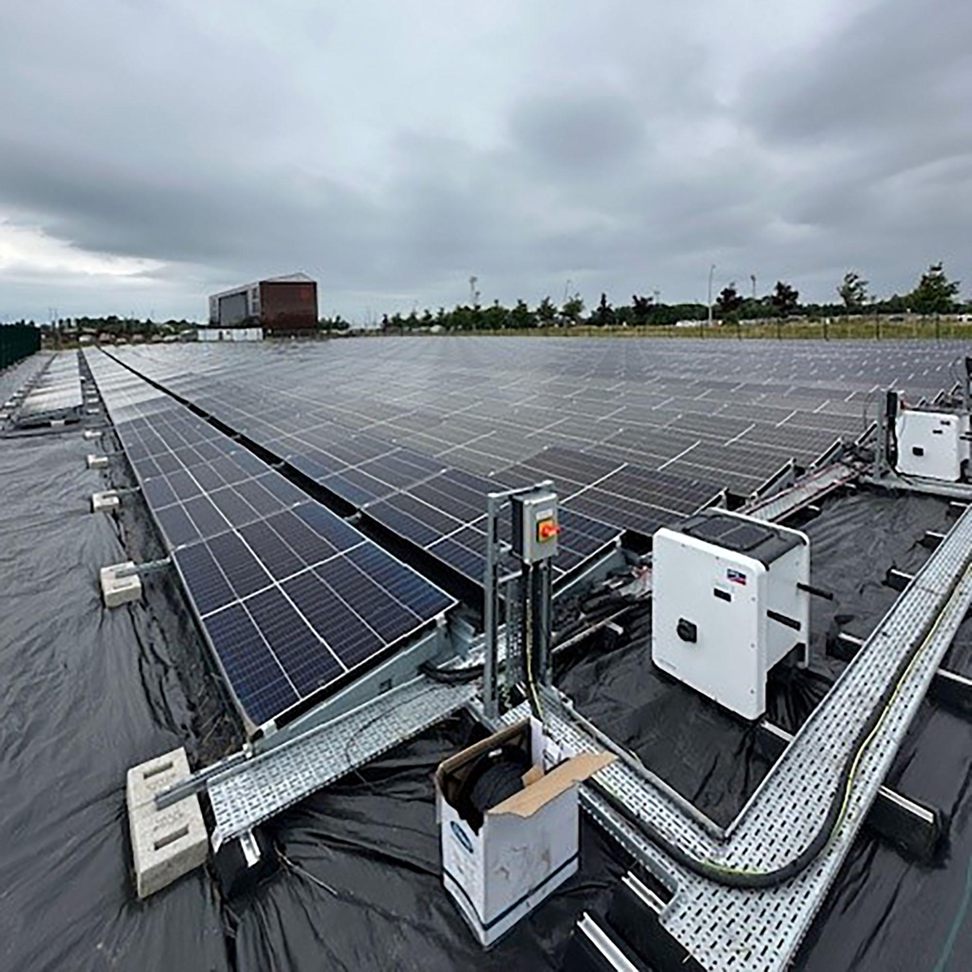 Rows of solar panels installed on a flat, black surface under a cloudy sky, with electrical boxes and cables visible in the foreground. Trees and buildings can be seen in the background.