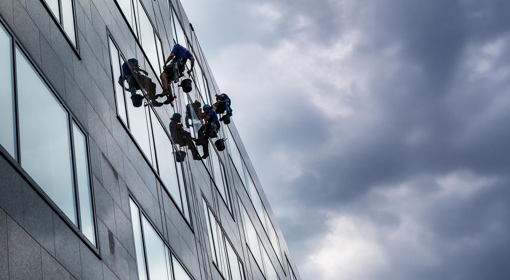 Three window cleaners, suspended by ropes, clean the windows of a tall building under a cloudy sky. Their reflections are visible in the glass.