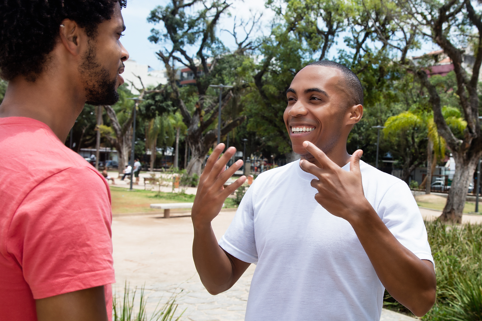 Two young black men talk with eachother outdoors near a park. One is using his hands to speak and smiling.