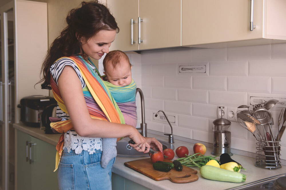 Mother with baby in a sling cuts vegetables in the kitchen
