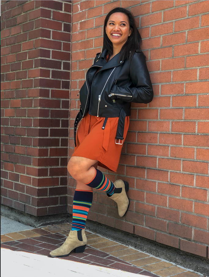Person leaning against brick wall wearing a leather jacket, orange dress, and blue and orange striped Dr. Segal's compression socks.