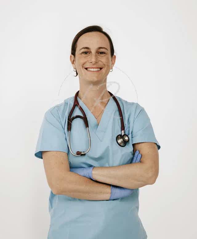 Smiling female doctor in light blue scrubs with stethoscope, arms crossed against a plain light background.