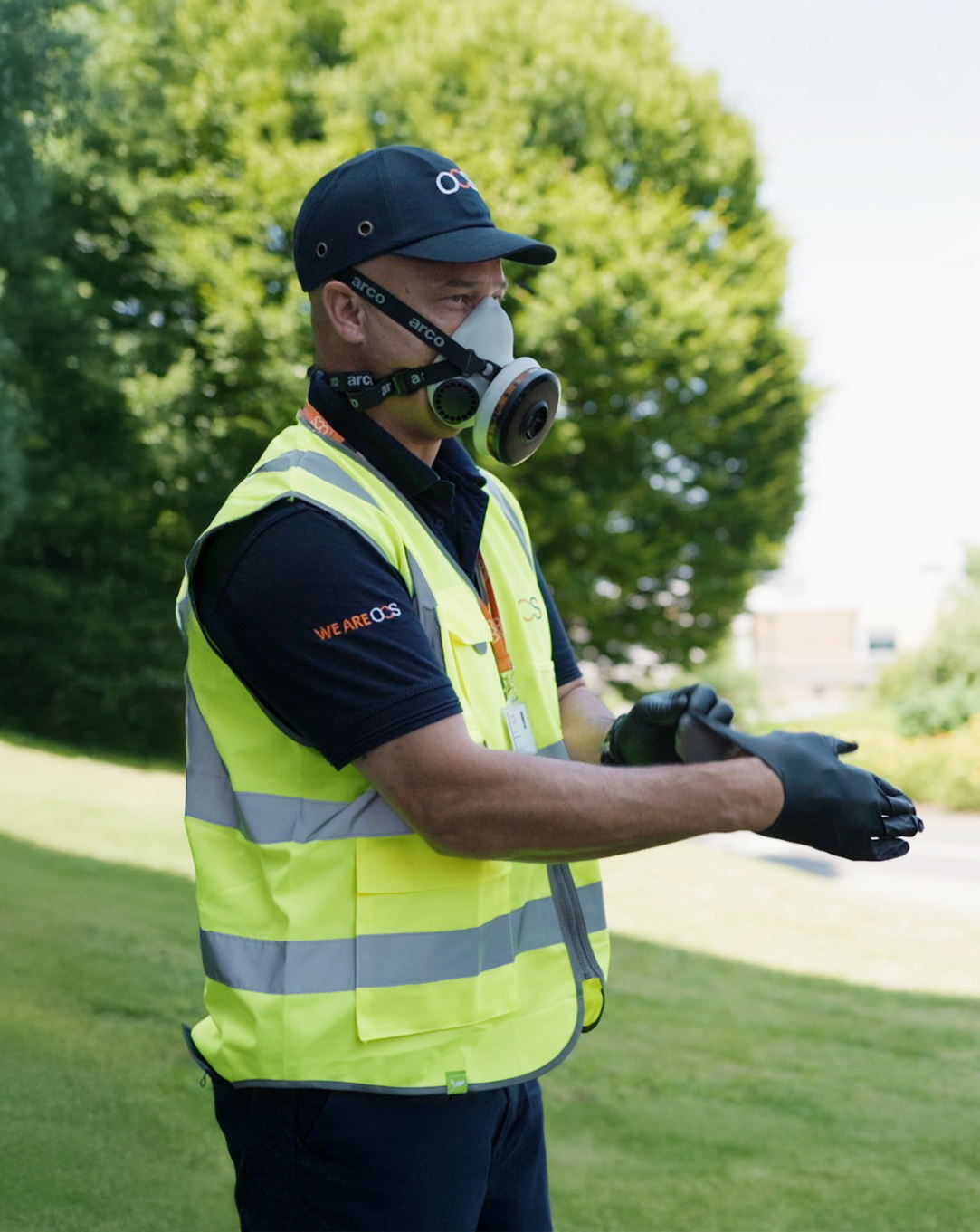 A person wearing a high-visibility vest, black gloves, a cap, and a face mask with a respirator stands outdoors on green grass, preparing to work. Lush green trees are visible in the background.