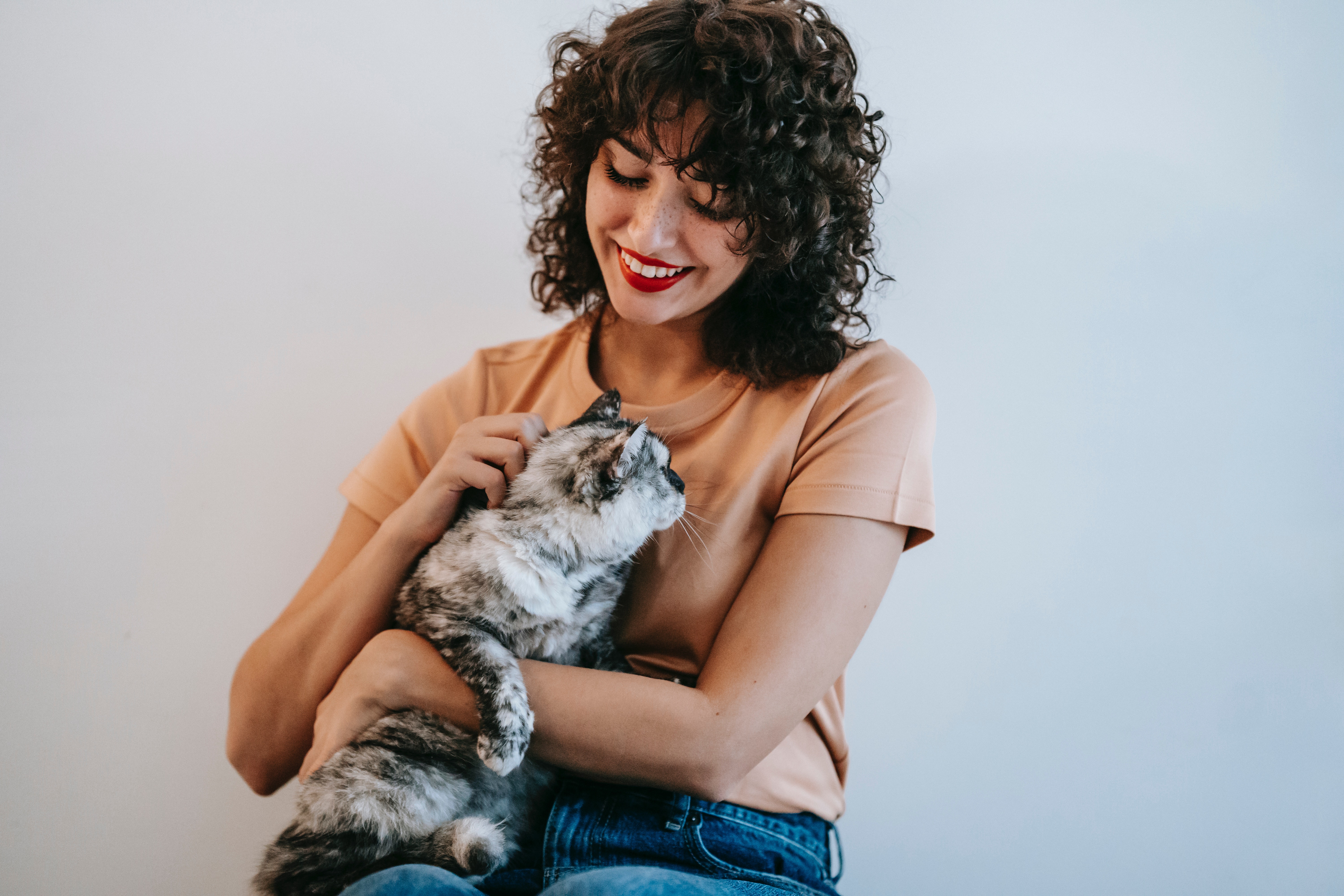 A woman with curly hair is sitting and looking at her cat in her lap and smiling to it.