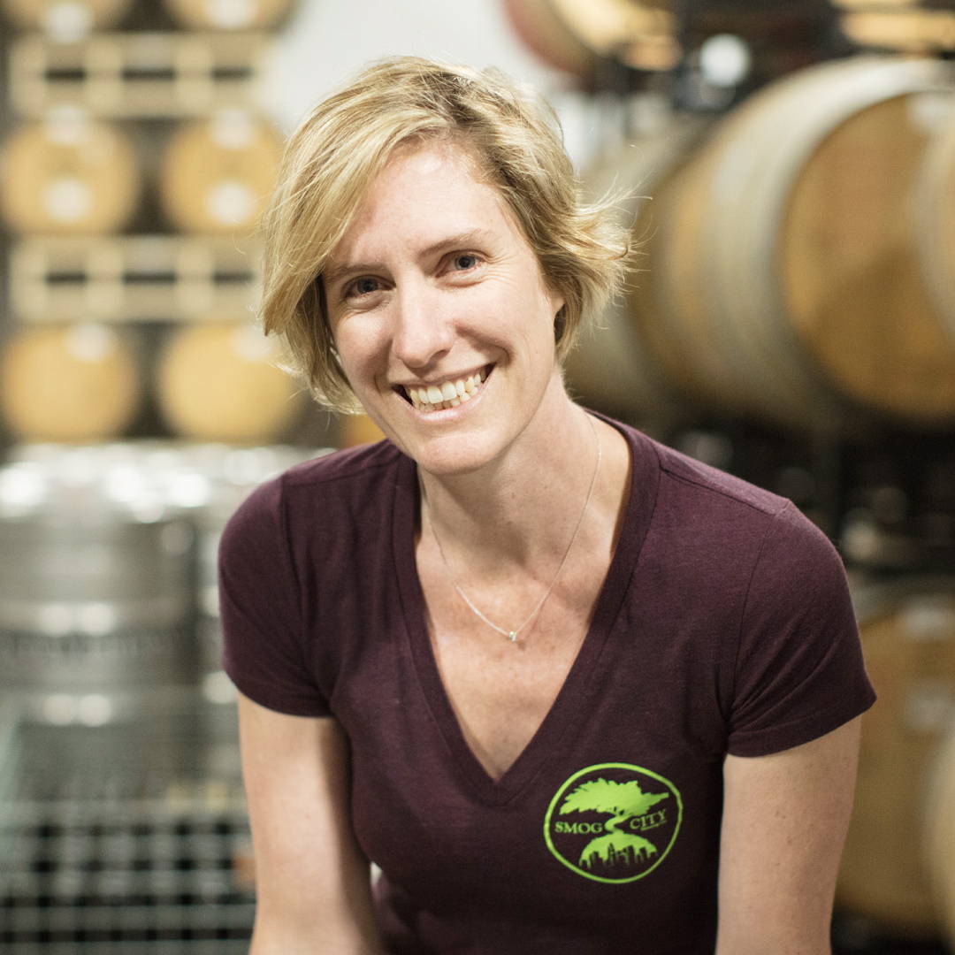 Owner, Laurie Porter, smiling in front of stacked wooden barrels and beer kegs.