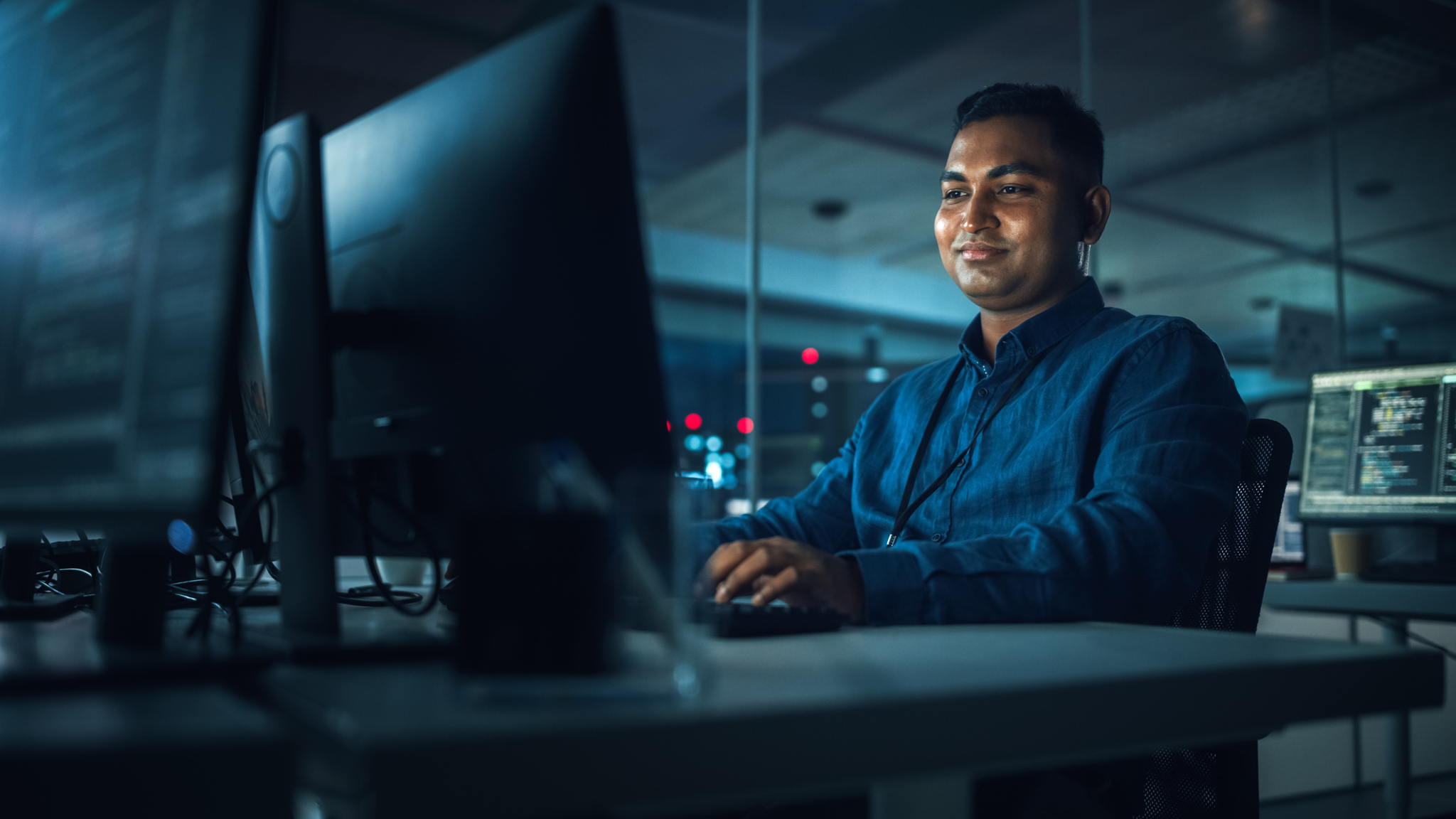 A man in a blue shirt sits at a desk in a dimly lit office, smiling as he works on a computer with multiple monitors displaying code and data.
