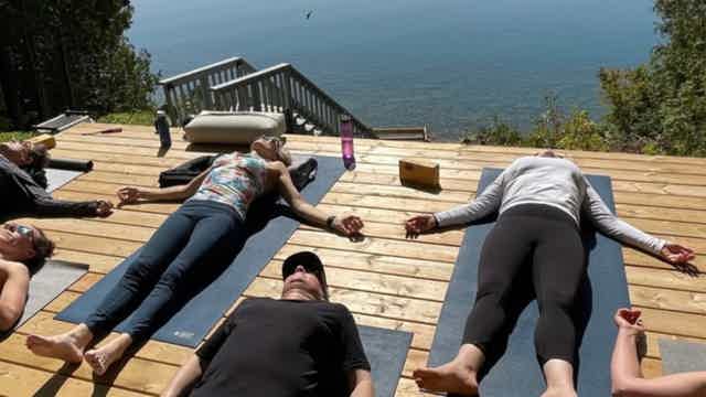 People lying on yoga mats on a wooden deck outdoors, overlooking water; some hold hands with others nearby.