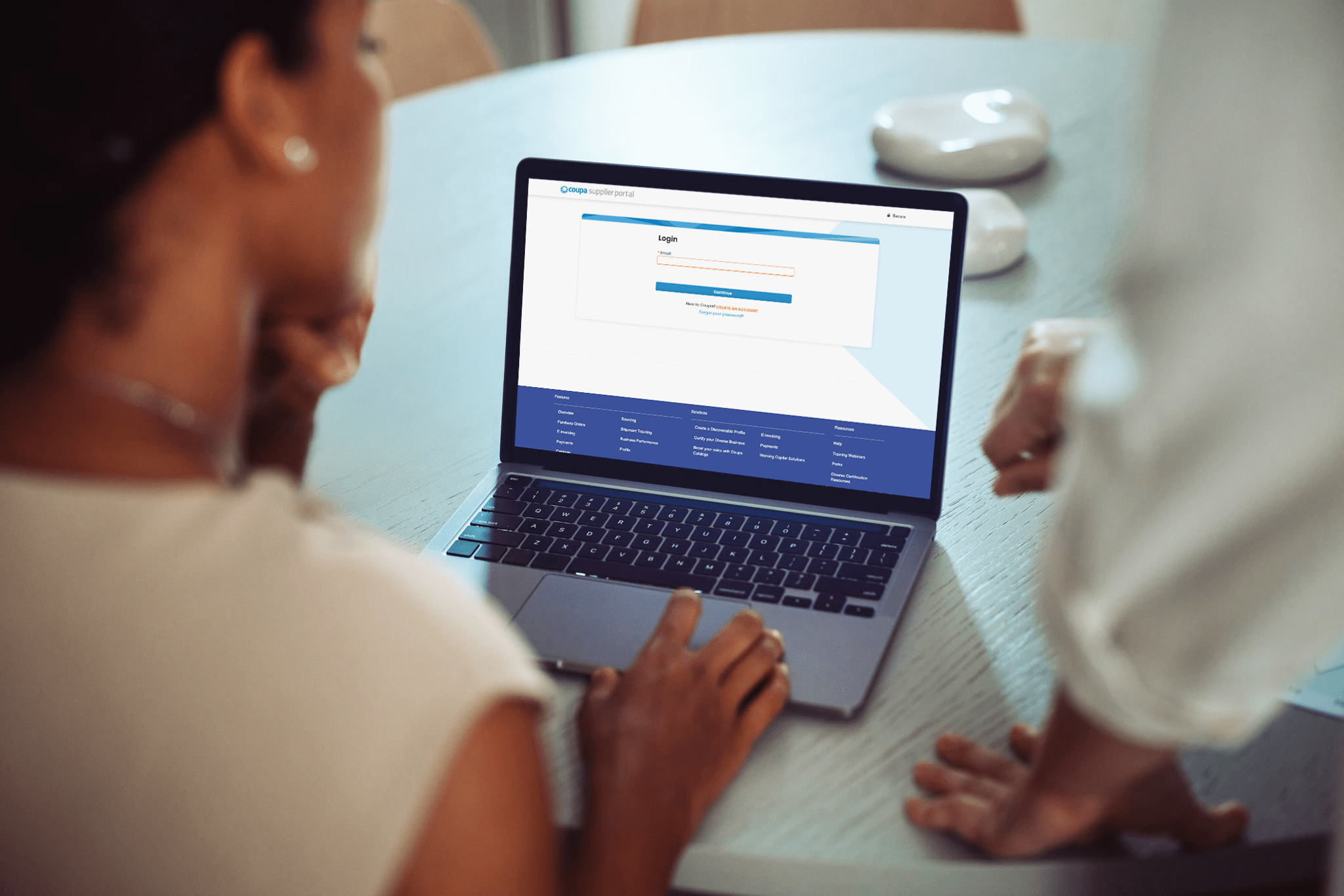 Two people are looking at a laptop screen displaying a login page for an online portal. One person is seated with a hand on the keyboard, while the other stands nearby. The focus is on the laptop screen.