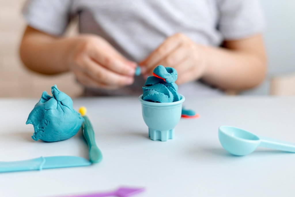 Un enfant sculptant avec de la pâte à modeler bleue, créant une forme de glace à l'aide d'ustensiles de jouet et d'une tasse de jeu sur une table blanche.