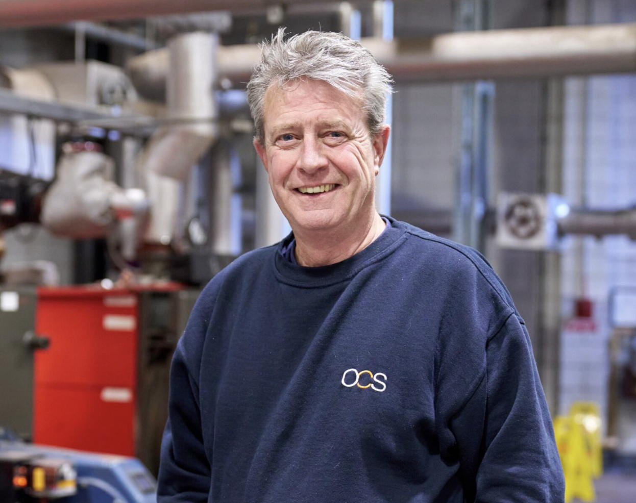 A middle-aged man with gray hair wearing a navy blue sweatshirt that says OCS stands smiling in an industrial setting with pipes and machinery in the background.
