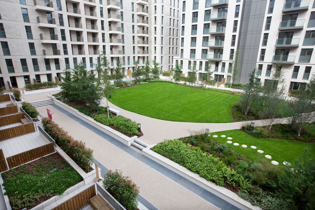 A modern apartment complex surrounds a landscaped courtyard with a large, oval-shaped green lawn, walking paths, trees, shrubs, and garden beds. Balconies overlook the courtyard from multiple floors.