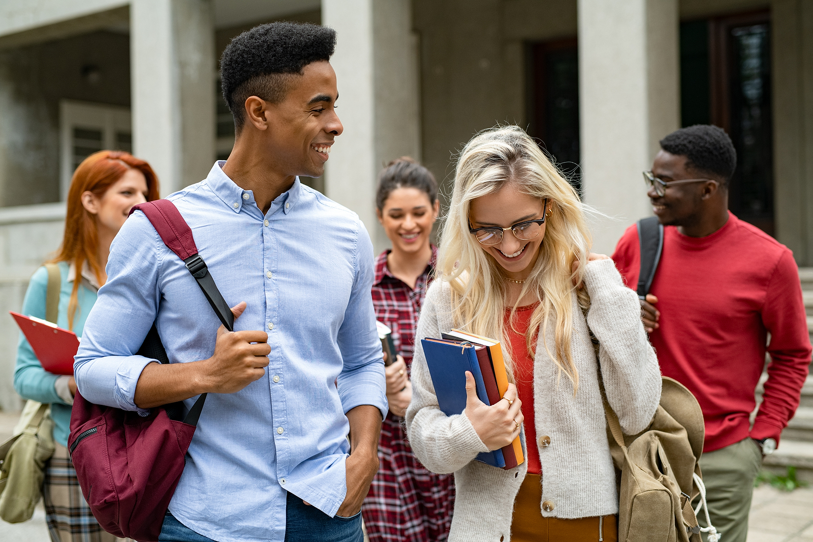 A group of young multi ethnic students walk to class with their backpacks. At the front is a guy and girl flirting with eachother, laughing together.