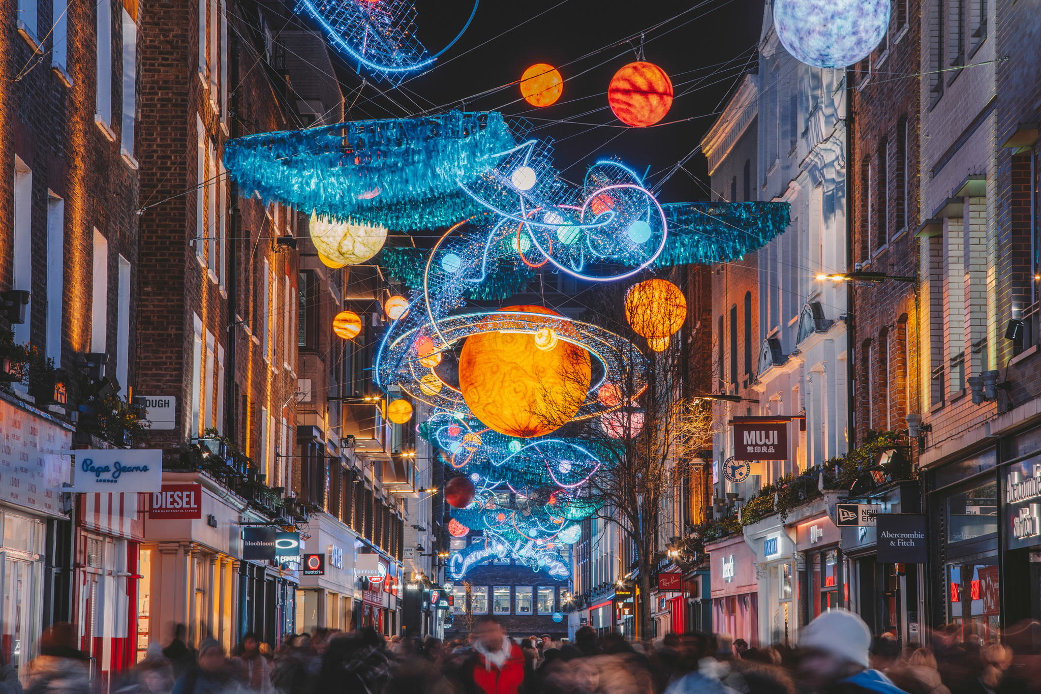 A busy city street at night is decorated with colorful, illuminated planets and orbital patterns hanging above, resembling a solar system, while crowds of people walk below and shops line both sides.