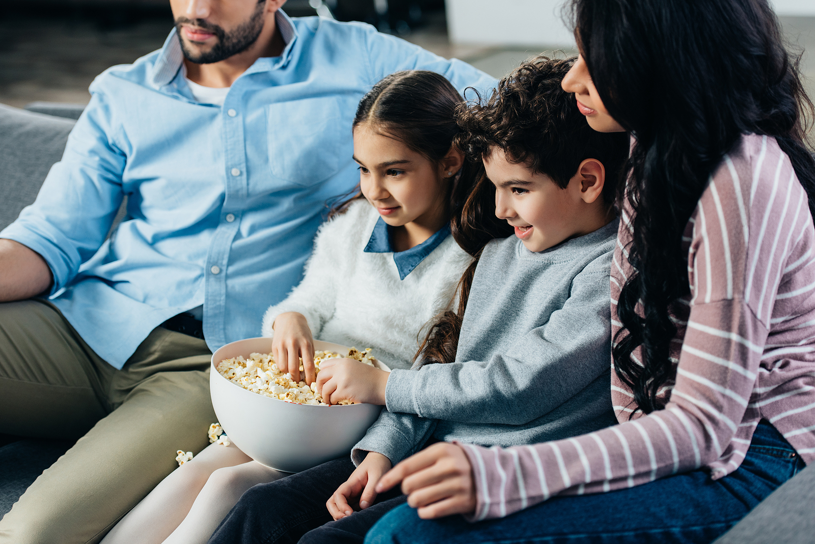 A nicely dressed multiethnic family of 4 with a small boy and girl in the middle, eating popcorn sitting on a couch with their mom and dad.