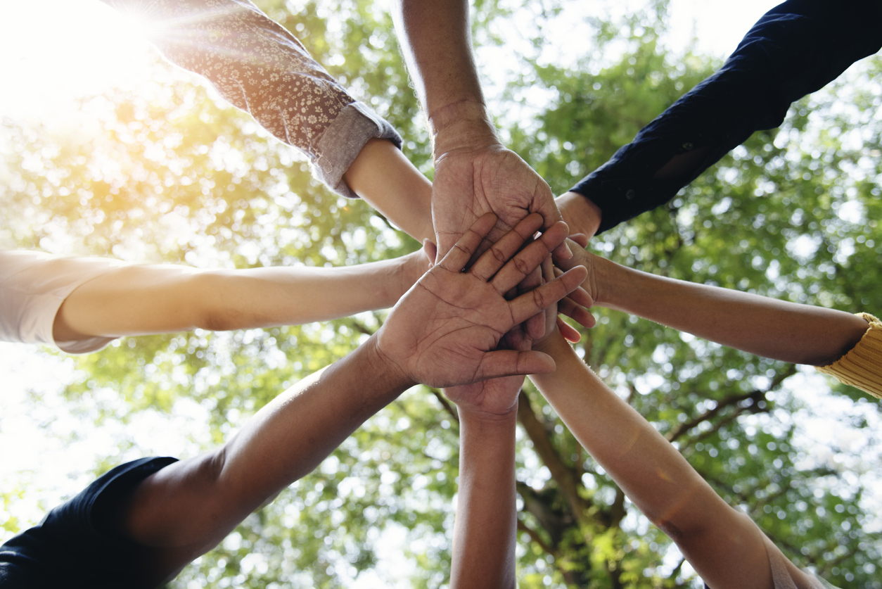 A group of people standing in a circle stack their hands together in the center, viewed from below, with sunlight and green trees in the background, symbolizing teamwork and unity.