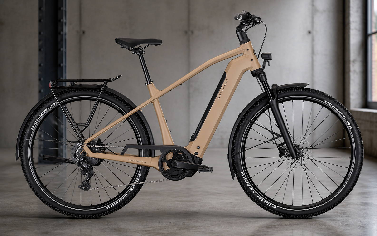 Beige Kalkhoff e-bike met zwarte onderdelen, bagagedrager en brede Schwalbe-banden, gefotografeerd in een industriële ruimte met betonnen vloer en grijze wand.