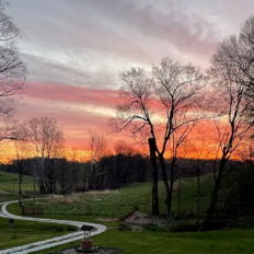 Sunset over a serene landscape with a winding path, lush green yard, and hills, evoking tranquility and reflection.