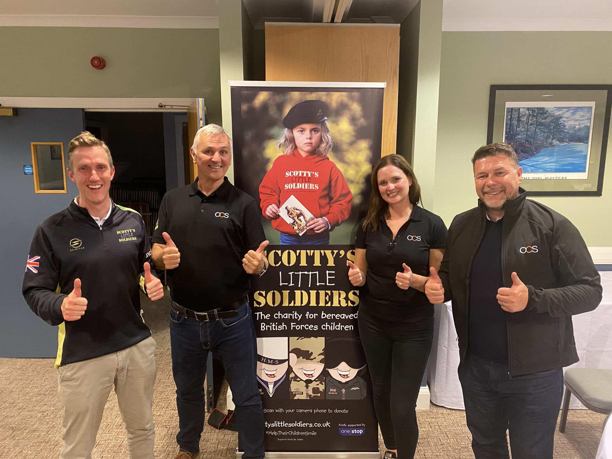 Four smiling adults stand indoors, giving thumbs up beside a Scottys Little Soldiers charity banner for bereaved British Forces children. They wear branded shirts and jackets and pose in a friendly group.