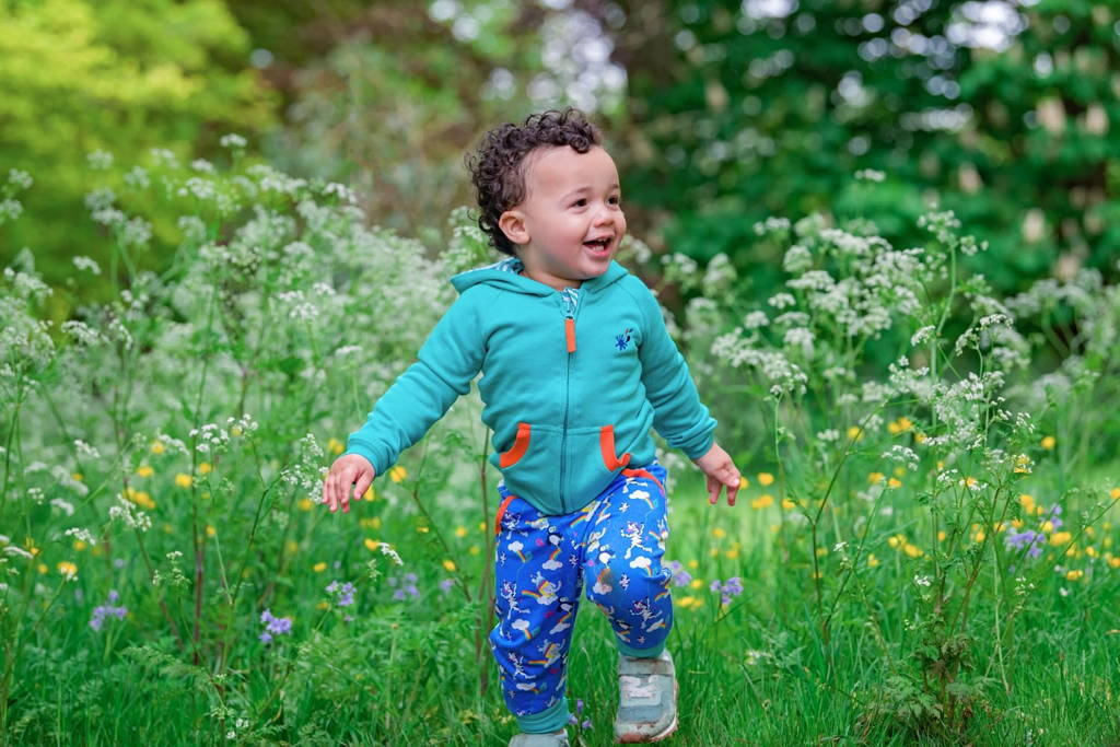 Image of a smiling toddler surrounded by long grass and wild flowers, wearing a Ducky Zebra turquoise hoody and brightly coloured Ducky Zebra trousers