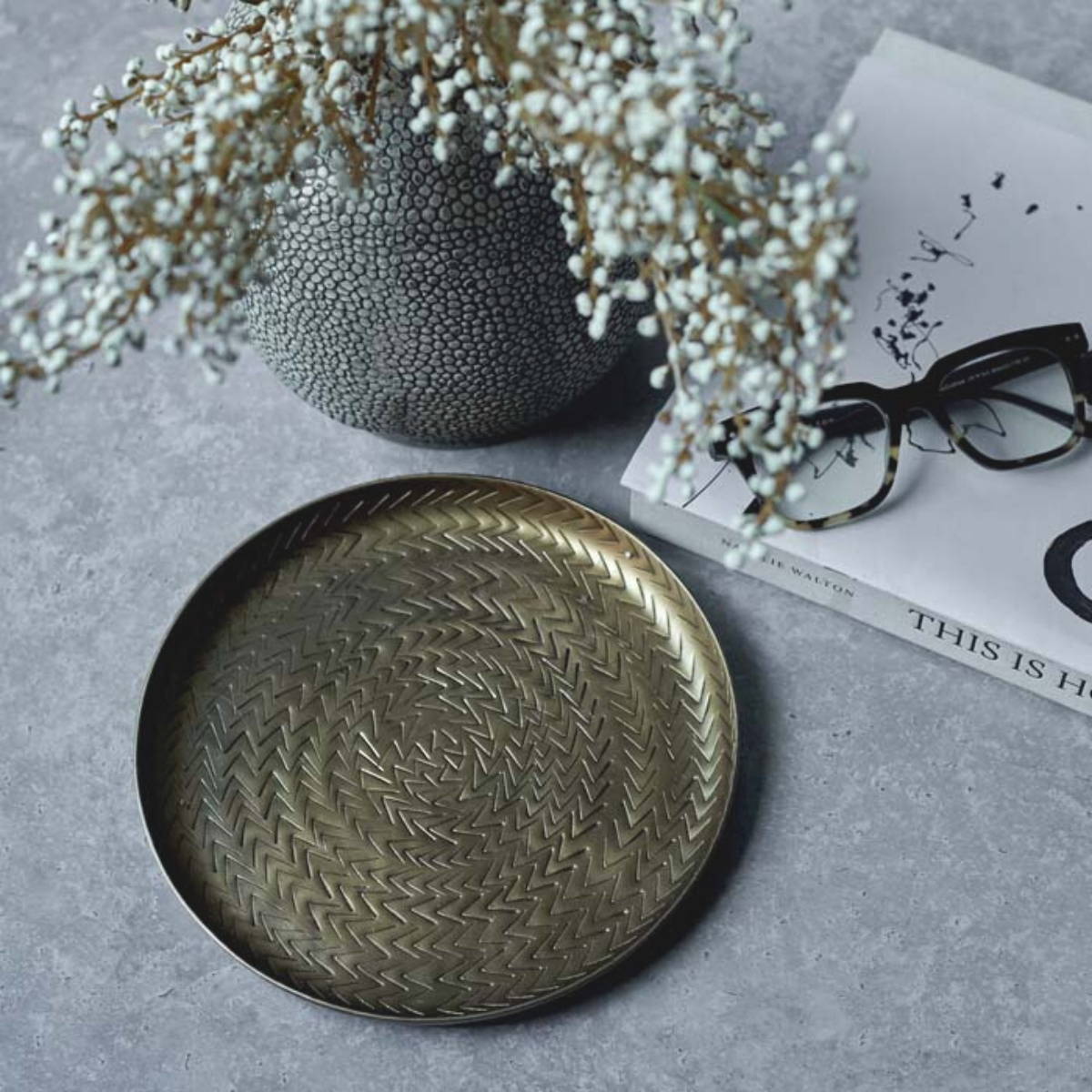 a small round brass tray with decorative detail next to a vase with some artificial flowers and some reading glasses and a book about home