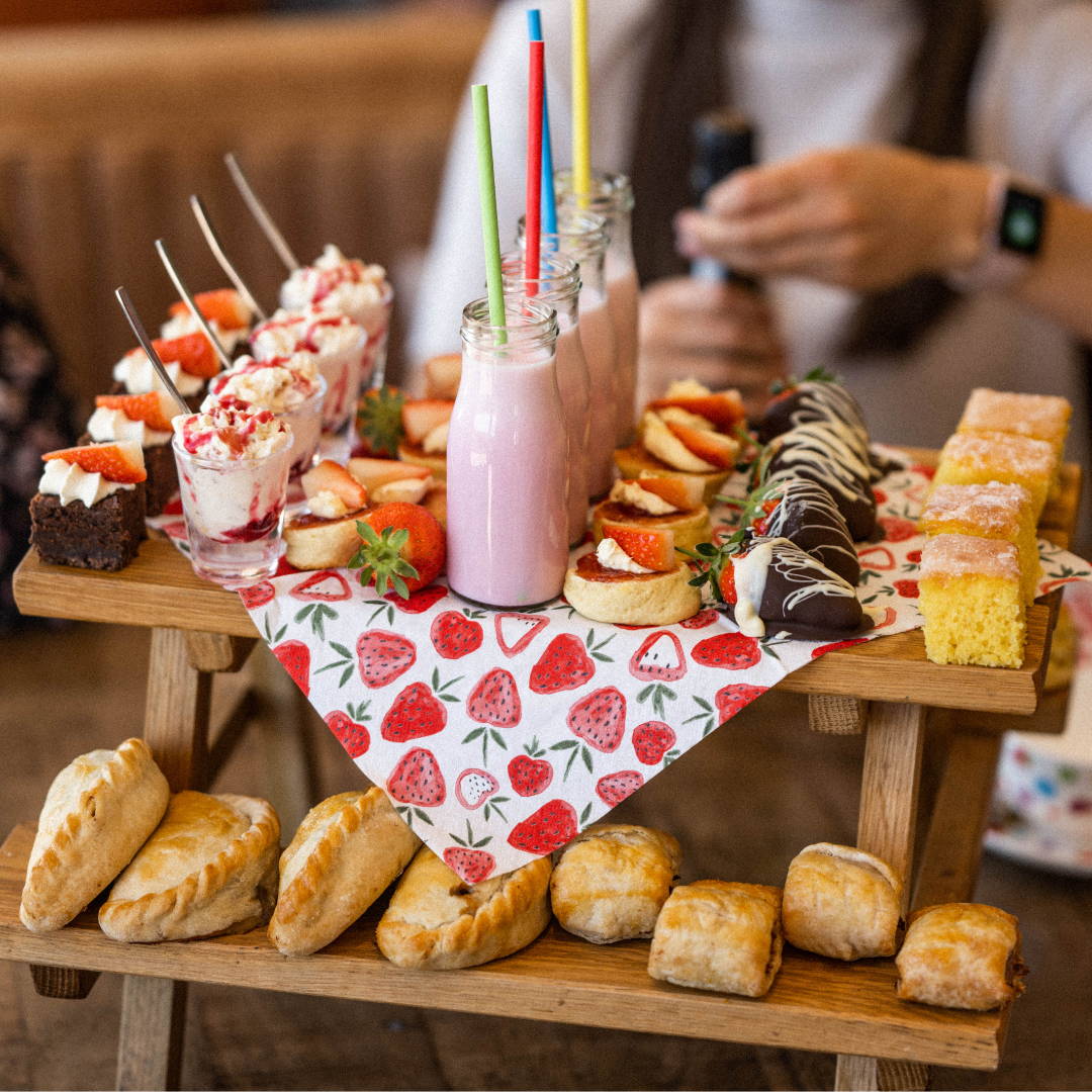 Afternoon tea, selection of strawberry milkshake, scones, cakes, pasties and sausage rolls displayed on a tiered wooden mini bench with a strawberry table cloth