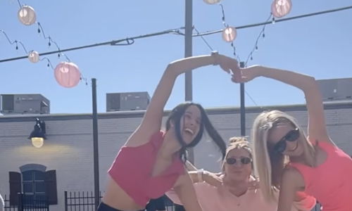 Three friends in matching pink outfits form heart shapes with their arms, smiling joyfully in a vibrant outdoor setting.