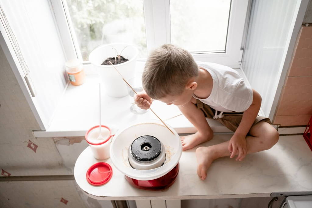Un jeune garçon assis sur un comptoir de cuisine faisant de la barbe à papa avec une machine à domicile près d'une fenêtre.