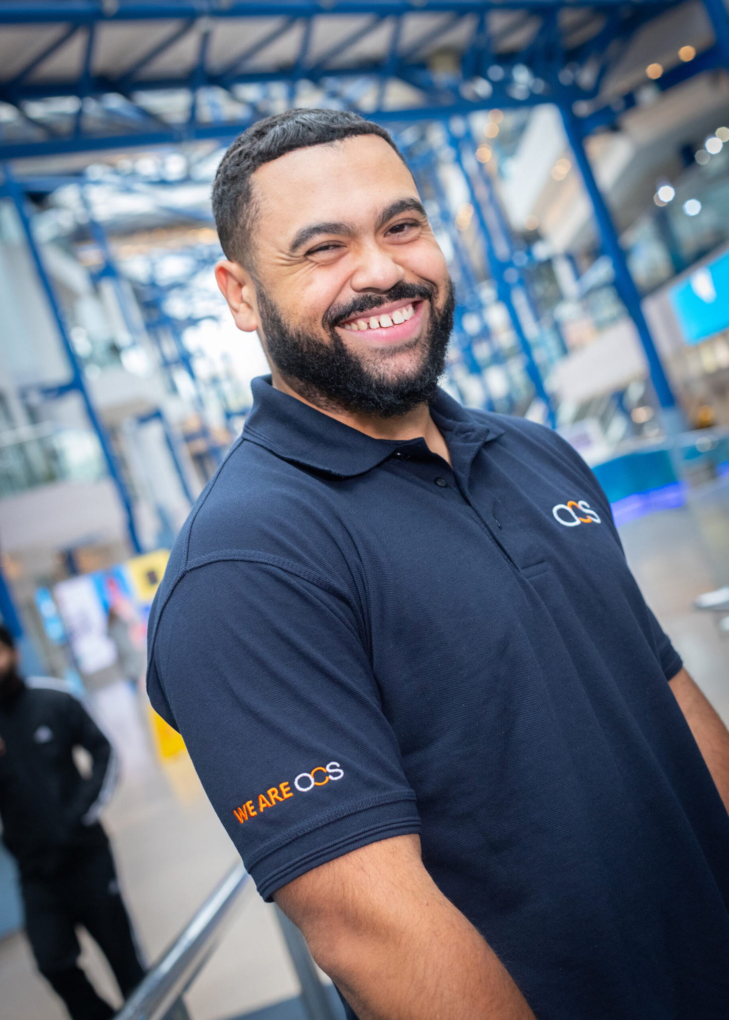 A smiling man with a short beard wearing a navy blue WE ARE OCS polo shirt stands indoors, with blue steel beams and bright lights in the background.
