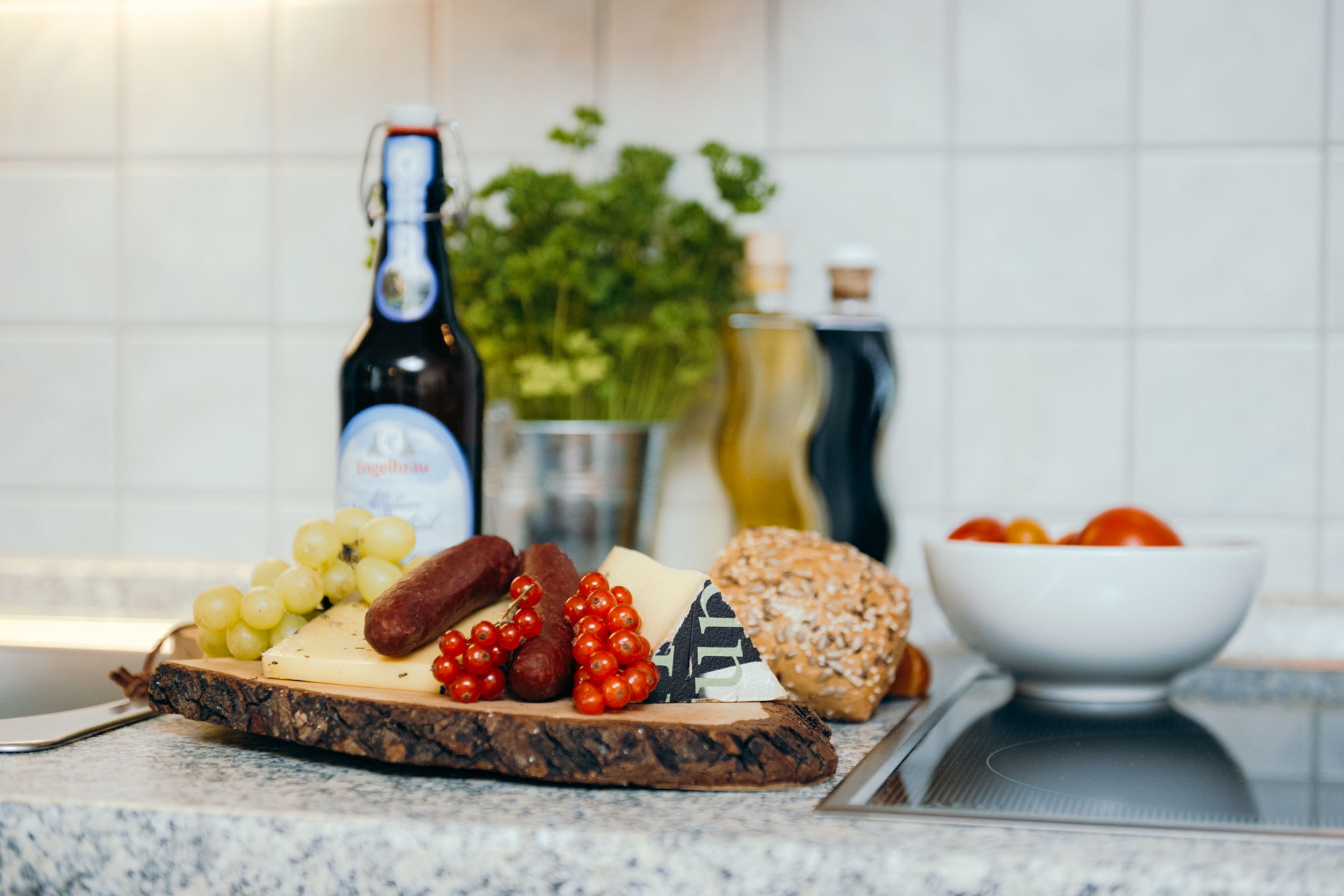 Auf einem Holzbrett liegen Brötchen, Käse, Wurst und Tomaten, im Hintergrund eine Schale mit Äpfeln, ein Bier, eine Basilikumpflanze und Ölflaschen.