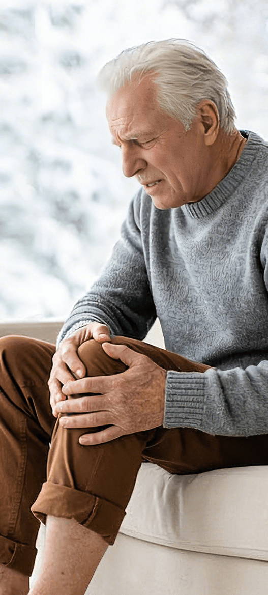 Older man sitting on a sofa holding his knee, showing mild arthritis or chronic joint pain in the knee