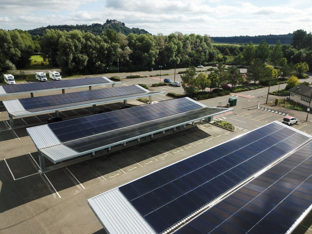 Rows of solar panels installed on carports in a parking lot, surrounded by trees and greenery, with a few cars parked and a hill visible in the background under a partly cloudy sky.