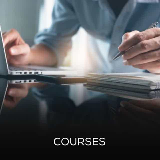 Two hands writing notes at a desk with stacked papers and a tablet, under soft light.